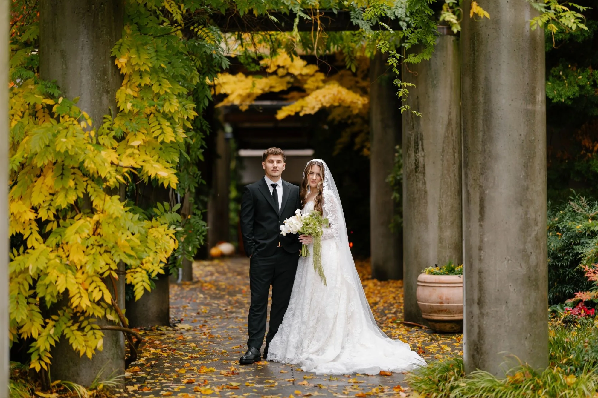 Bride and groom under a vine-covered pergola with golden autumn leaves