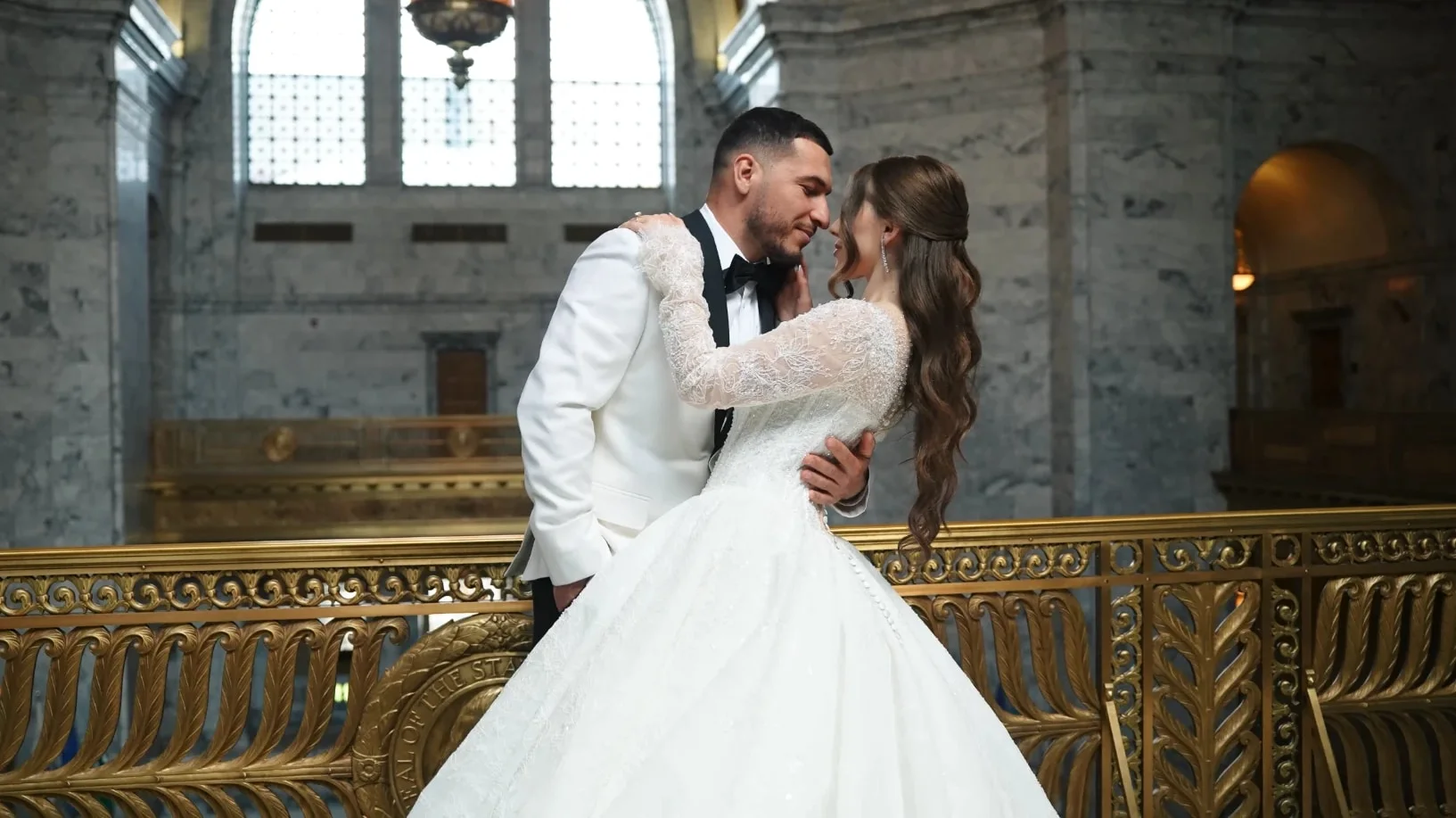Groom in a white tuxedo and his bride by a gilded railing in a marble hall