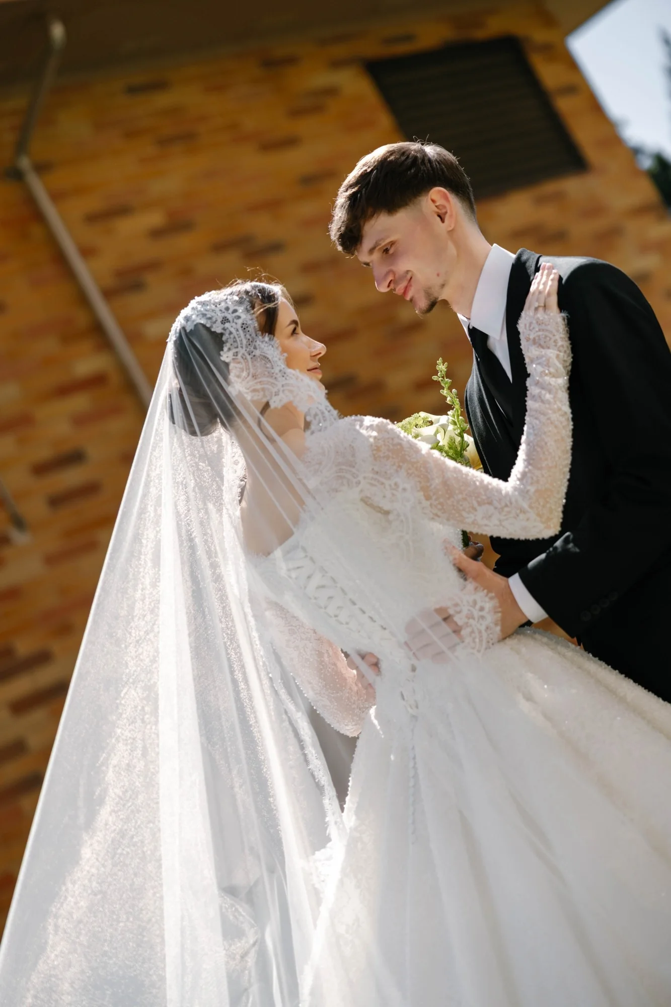Bride in a lace cathedral veil gazing up at the groom in the sunlight